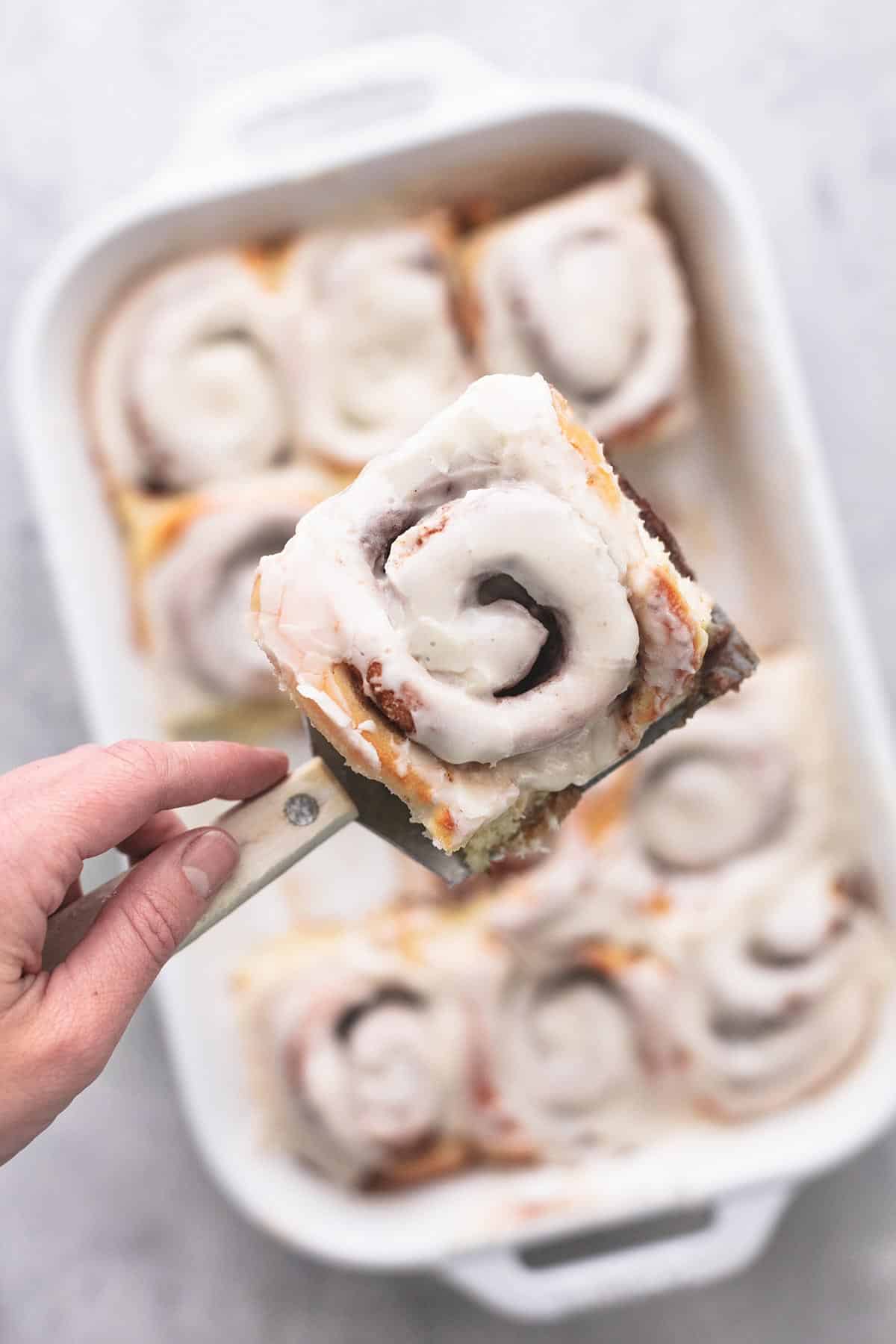 hand holding spatula holding single cinnamon roll above pan of more