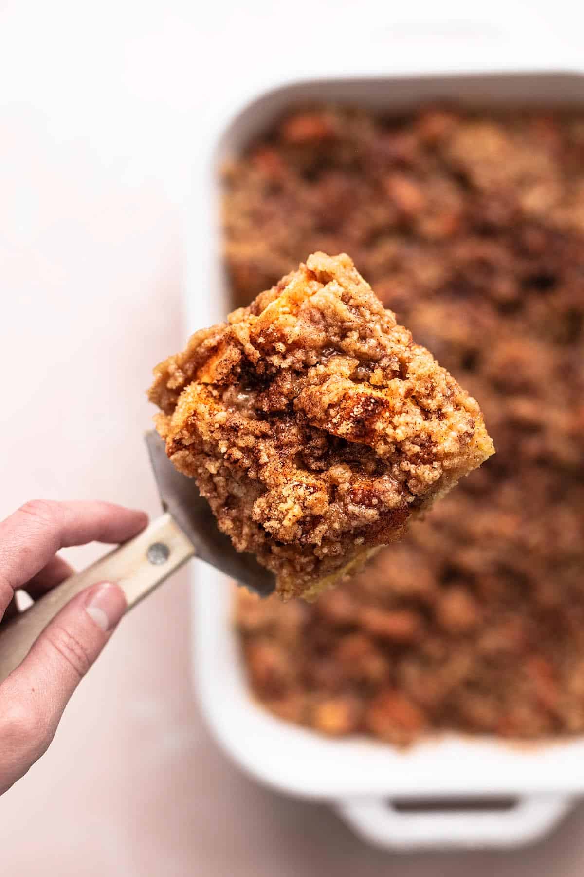 top view of a hand holding a spatula with a slice of french toast casserole above pan of more casserole.