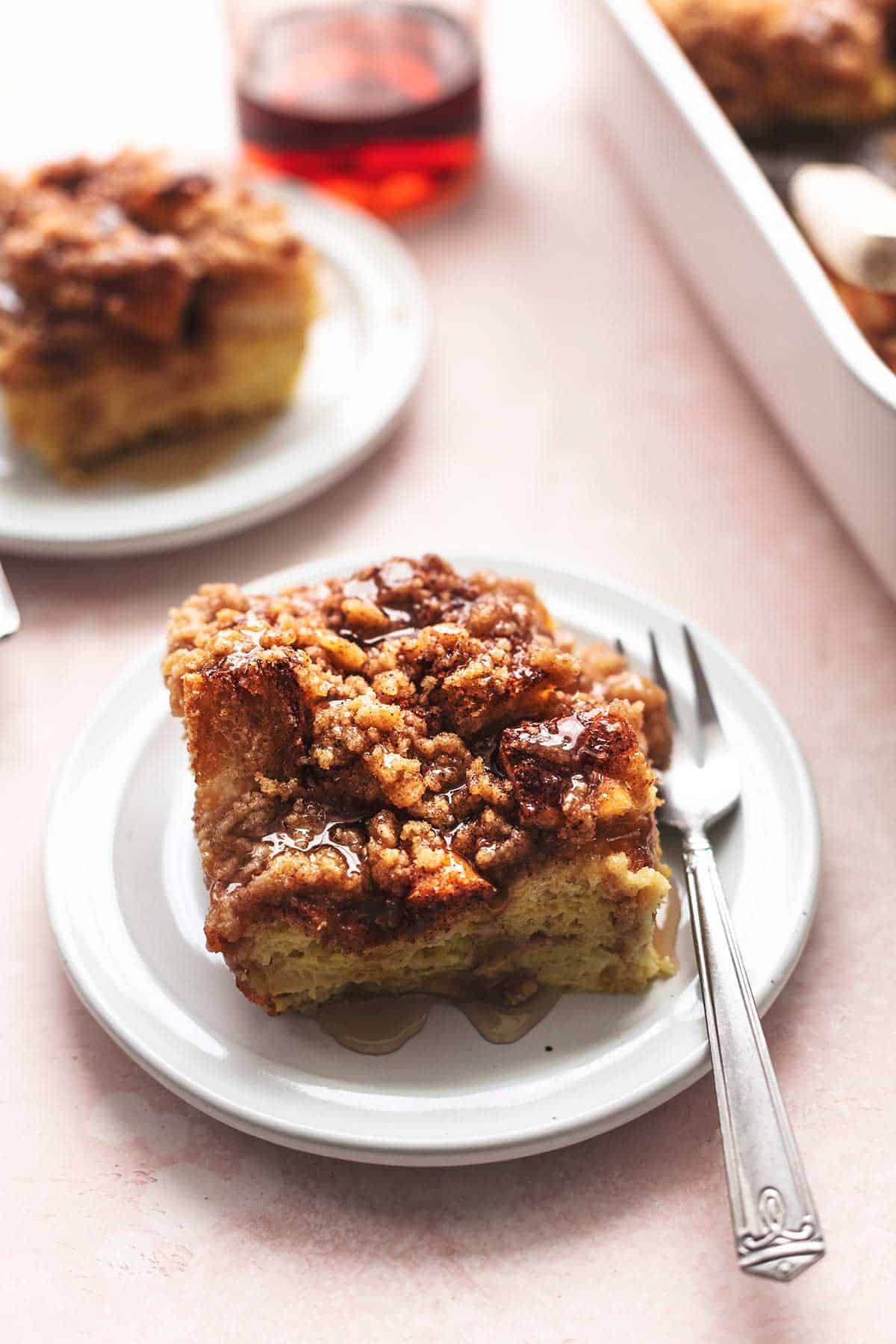 a piece of French toast casserole with a fork on a plate with another piece of casserole, a bottle of syrup and a baking pan of casserole in the background.
