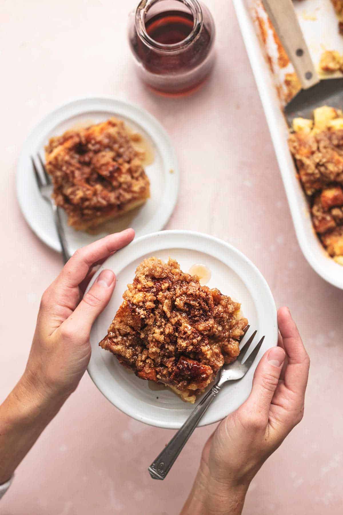 top view of hands holding a plate of French toast casserole with another plate, syrup, and a baking pan on the side.