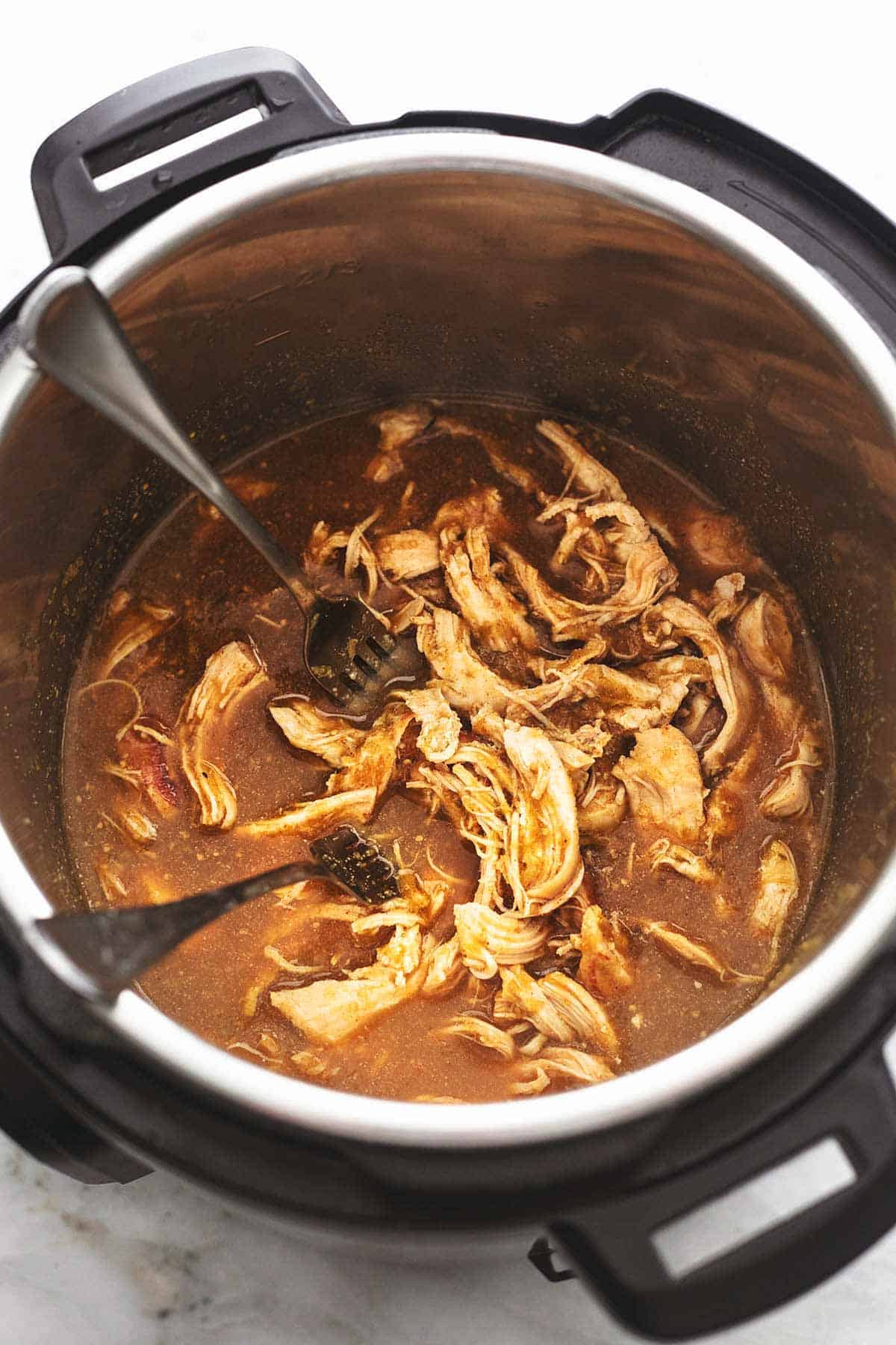 top view of shredded chicken with two forks in brown liquid inside a slow cooker.