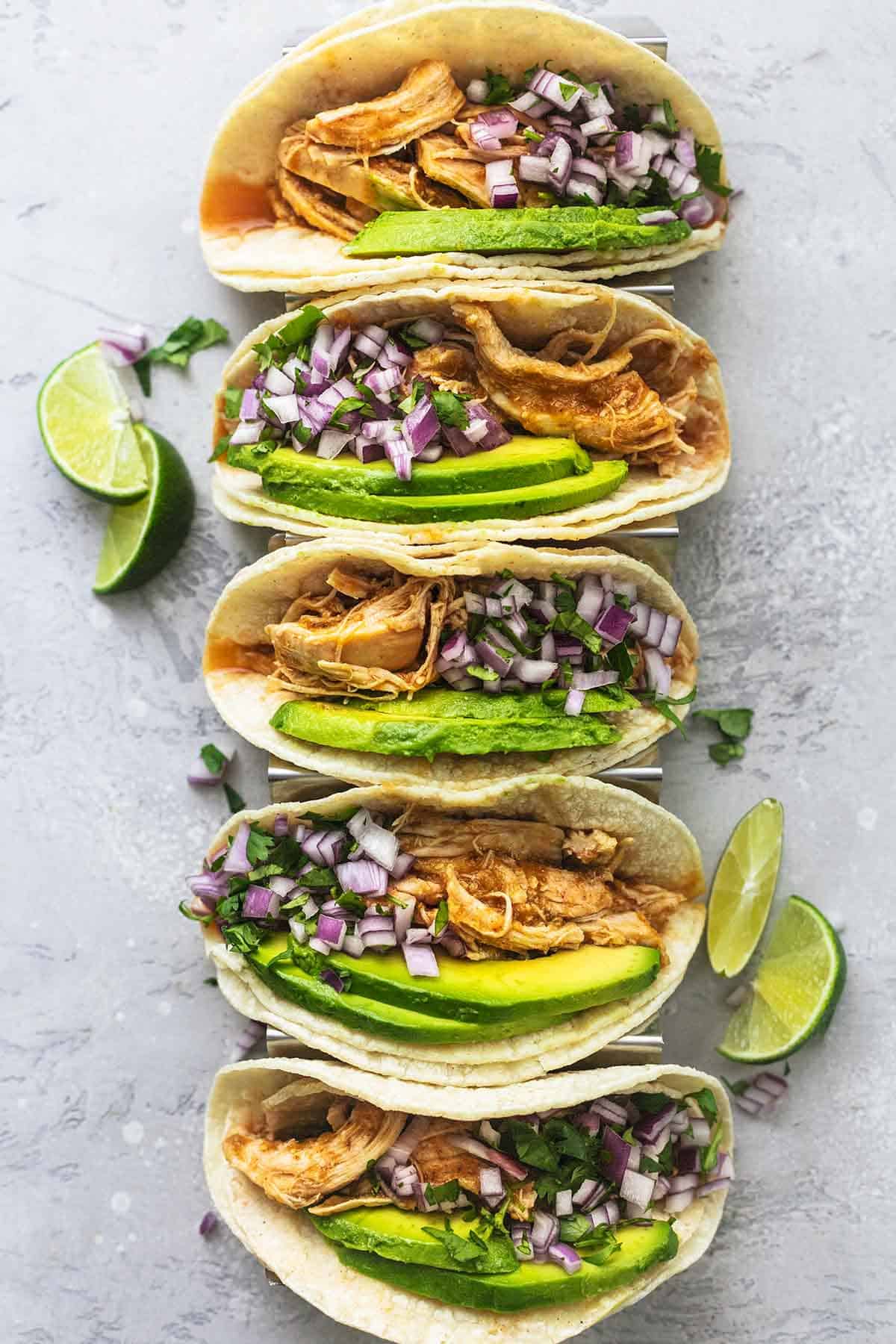 overhead view of five tacos with avocado and shredded chicken and salsa with limes on the side