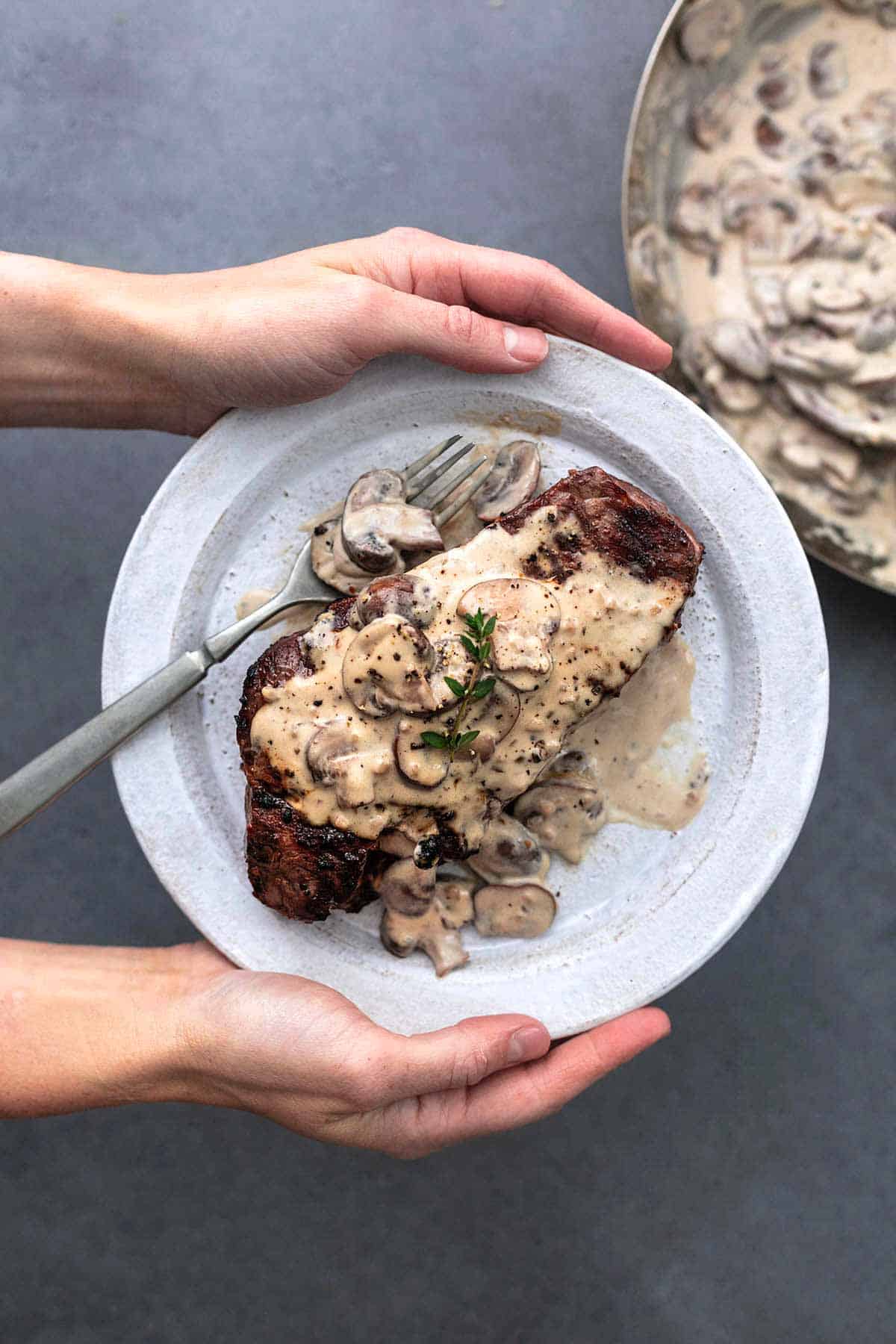 hands holding white plate with cooked steak topped with a brown mushroom sauce