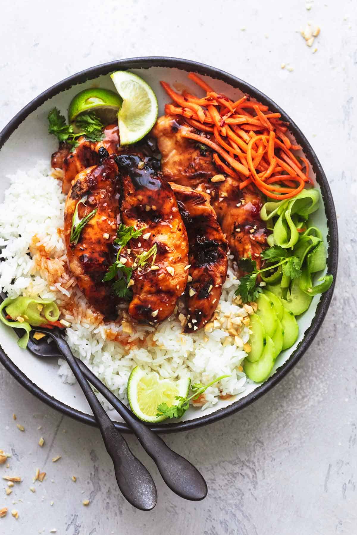 top view of sweet and spicy Thai chicken, rice, julienned carrots, cucumber ribbons, and lime wedges with forks on a plate.