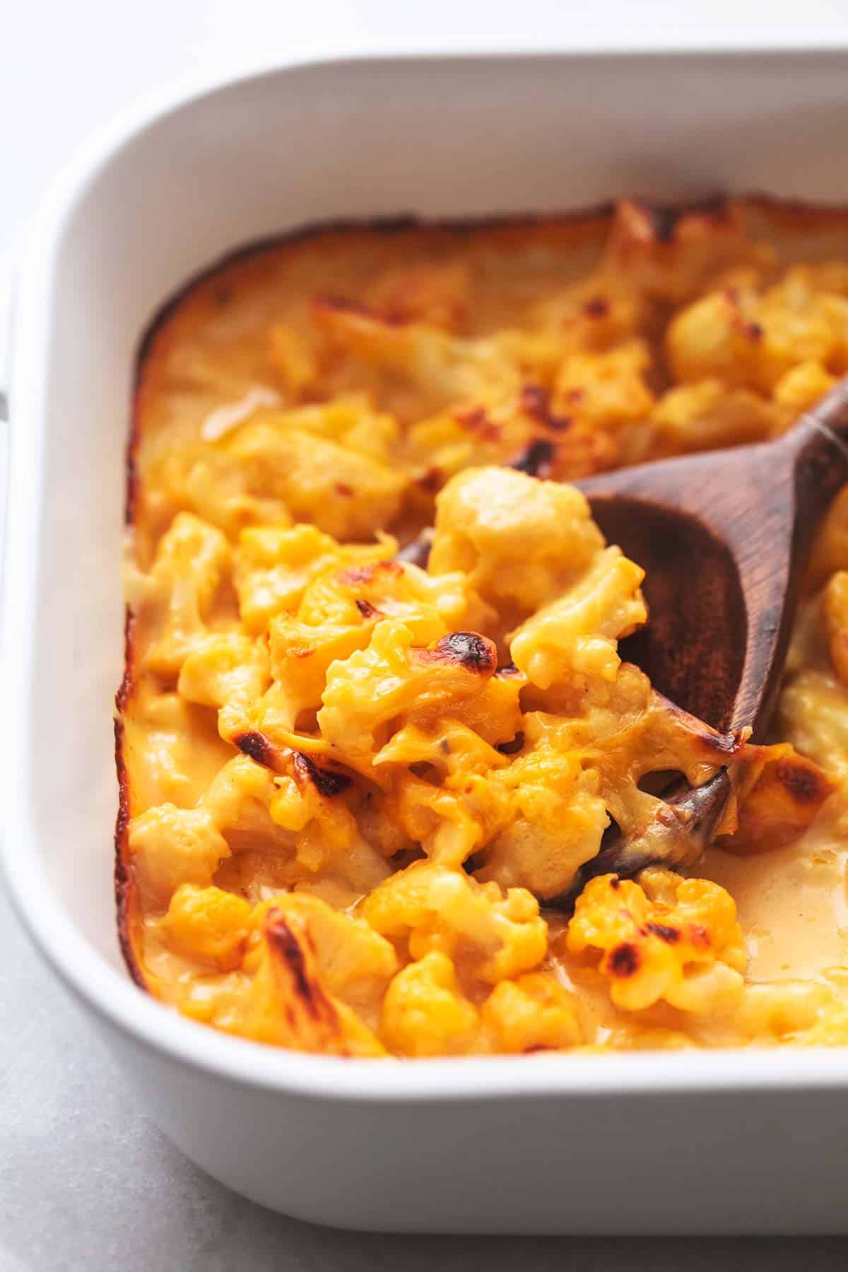 close up of baked cauliflower mac and cheese with a wooden serving spoon in a baking pan.