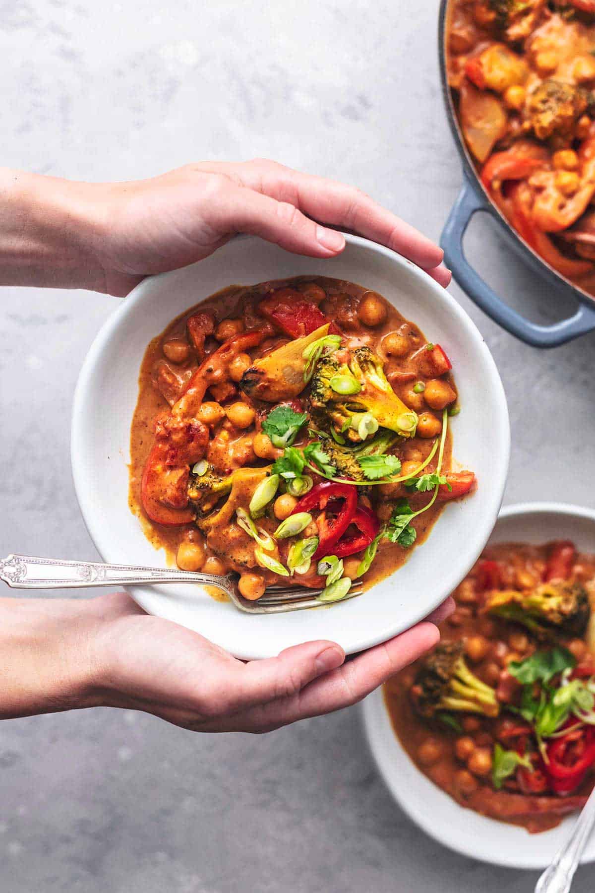 overhead view of hands holding bowl of chickpea curry with vegetables above second bowl and skillet