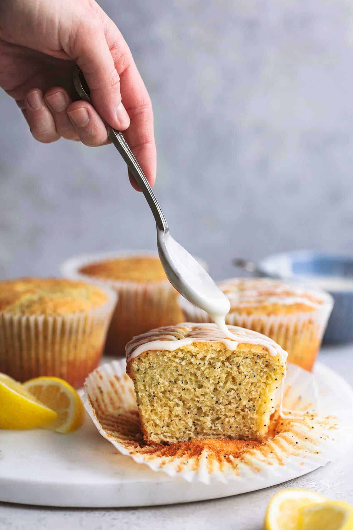 a hand holding a spoon drizzling white glaze on top of a lemon poppyseed muffin.