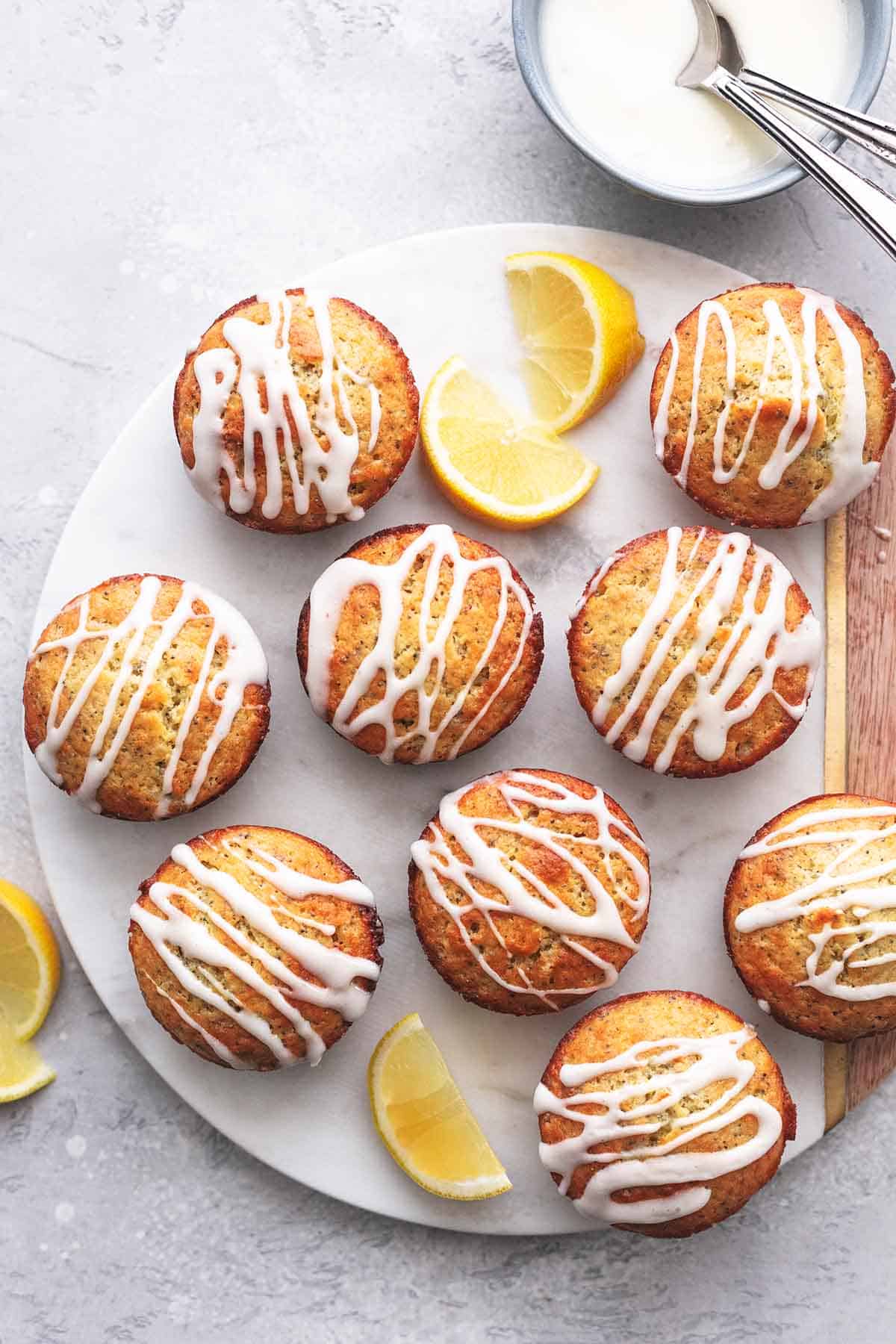 top view of lemon poppyseed muffins and lemon slices on a round cutting board with glaze in a bowl on the side.