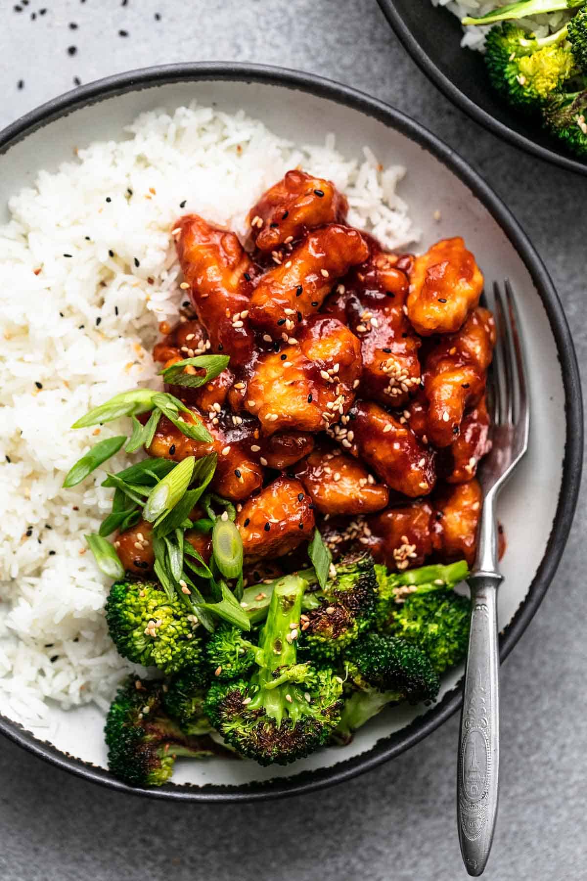close up top view of sticky sesame chicken with rice and veggies on a plate with a fork.