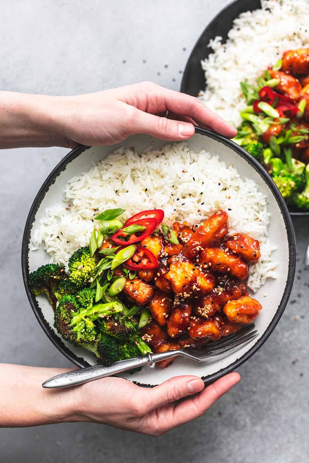 hands holding plate of sticky chicken over second bowl of food