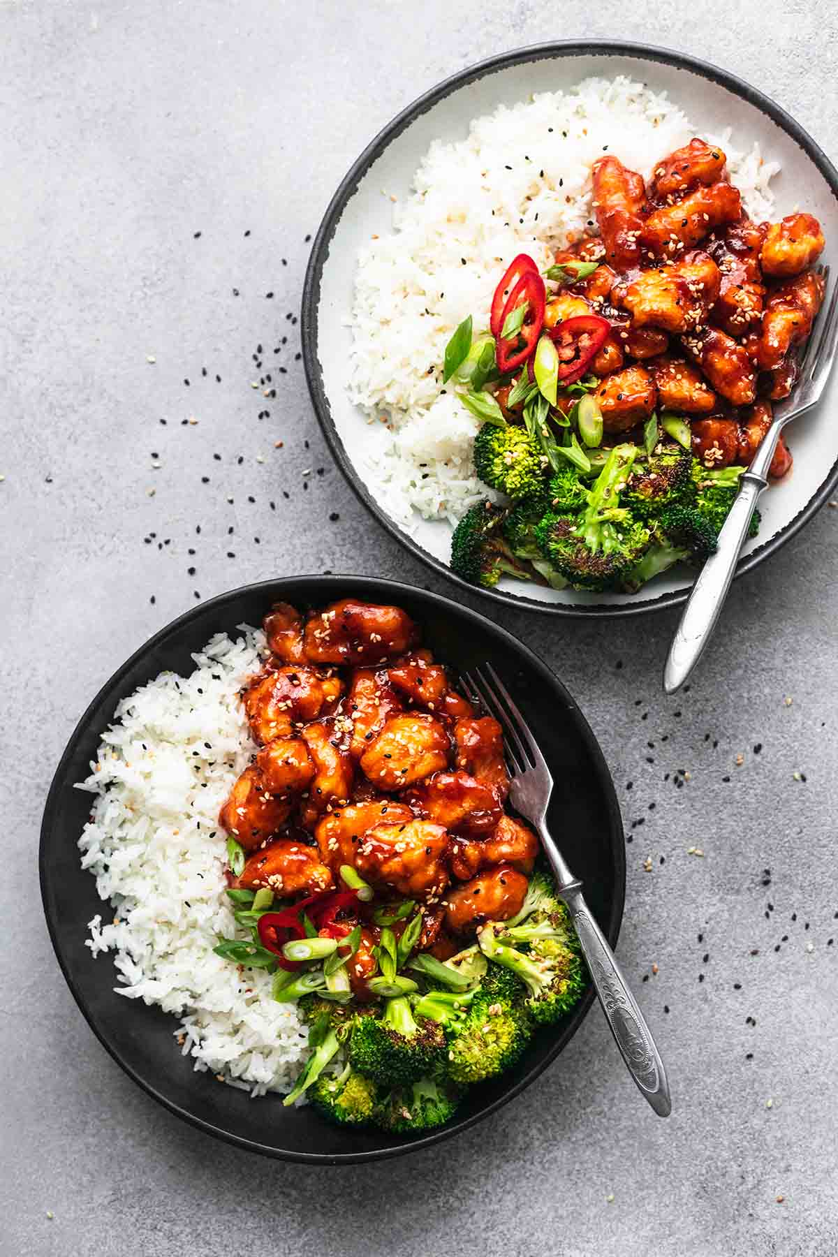 overhead view of two plates of asian chicken with sesame seeds and sides of broccoli and rice