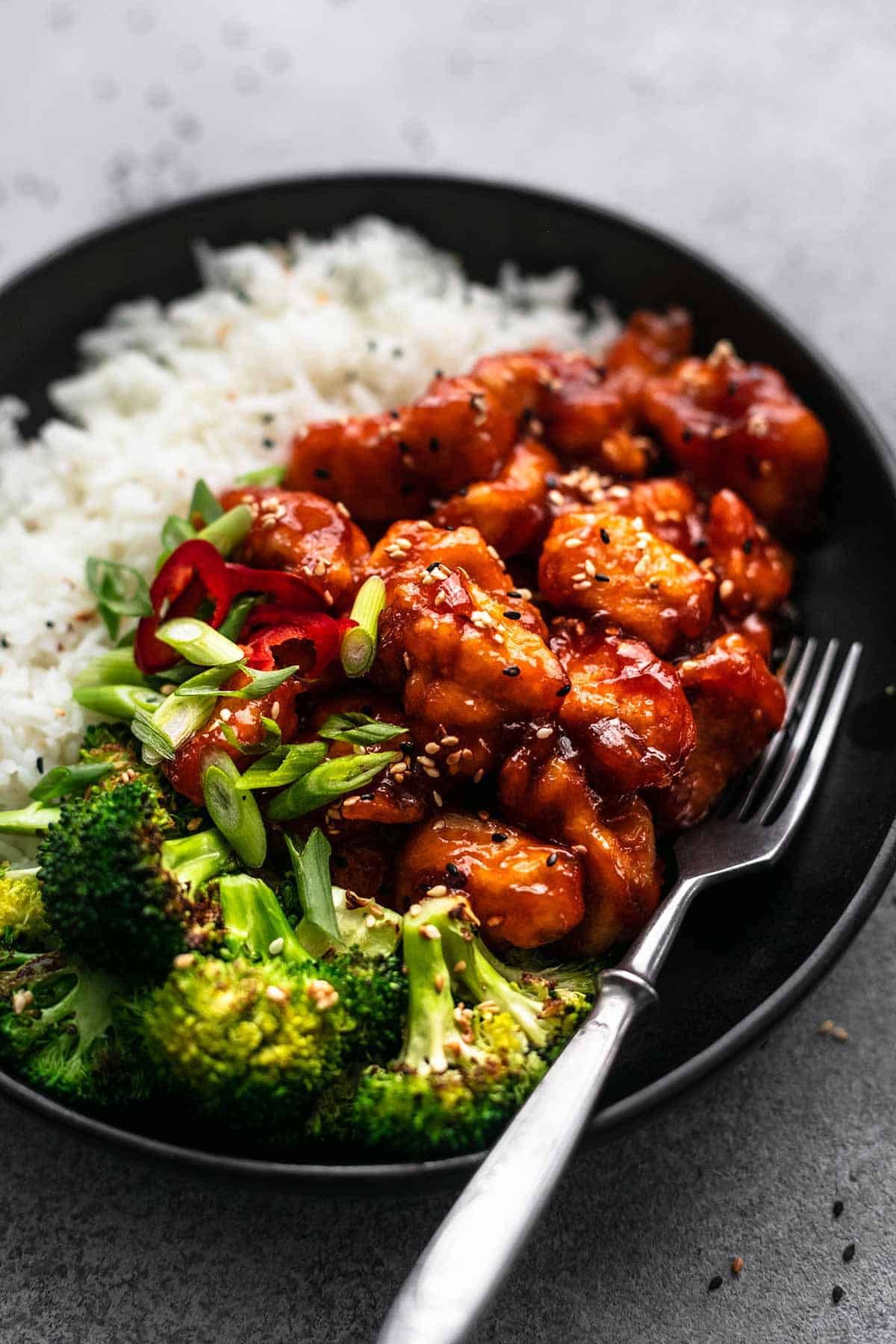 close up of sticky sesame chicken with rice and broccoli on a plate with a fork.