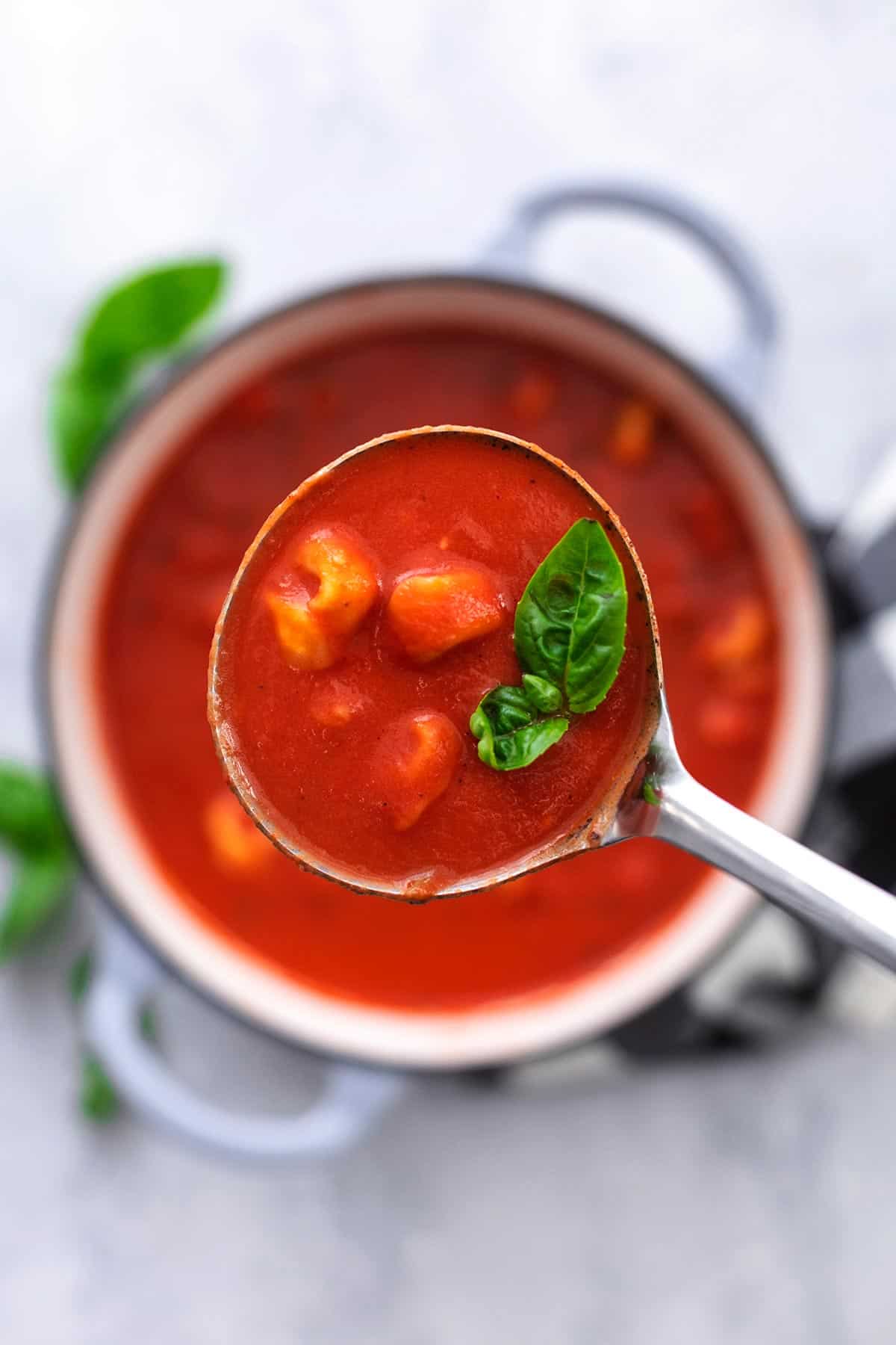 overhead view of ladle full of tomato soup with tortellini above pot of more soup