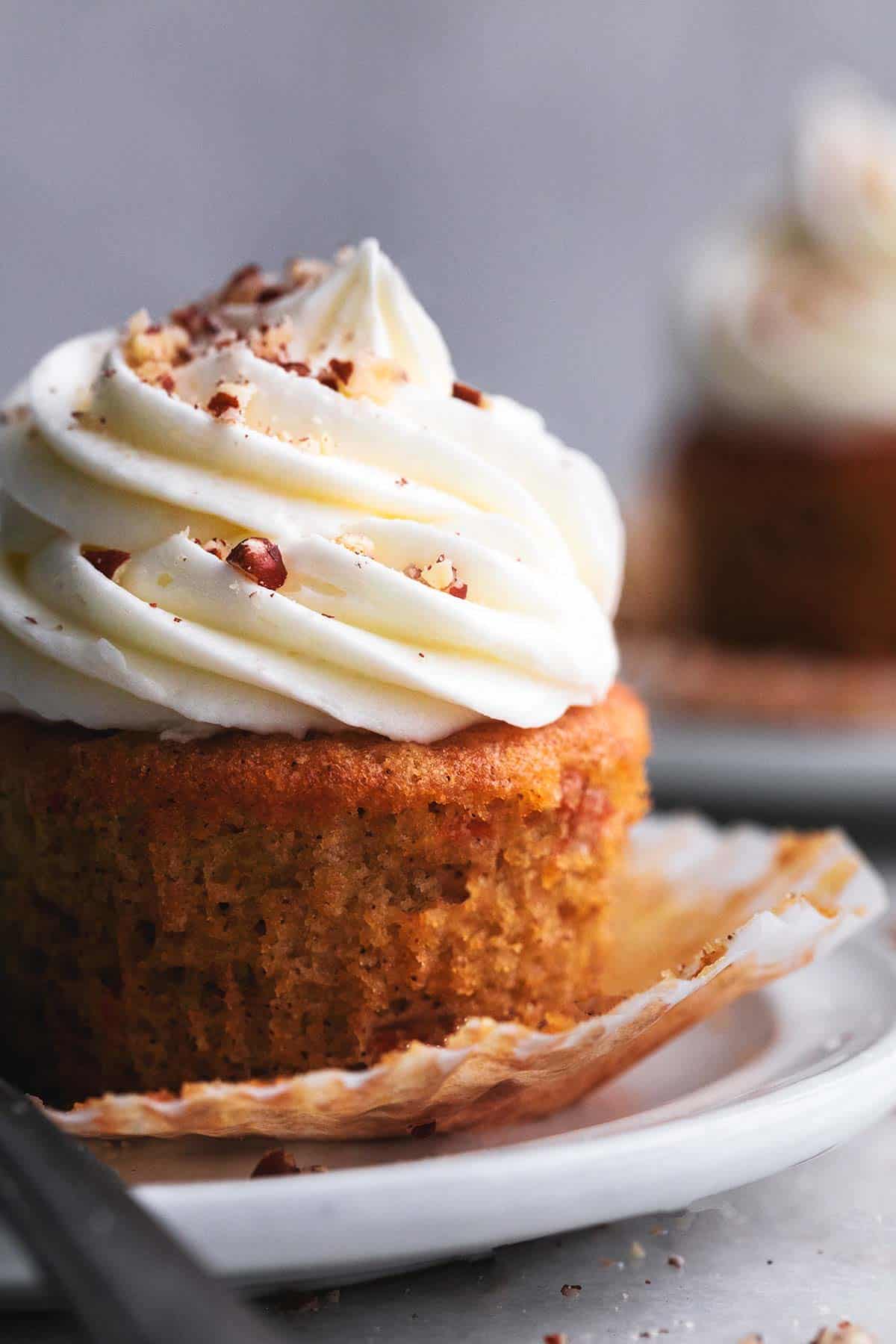close up of a carrot cake cupcake with cream cheese frosting with the liner open on a plate.