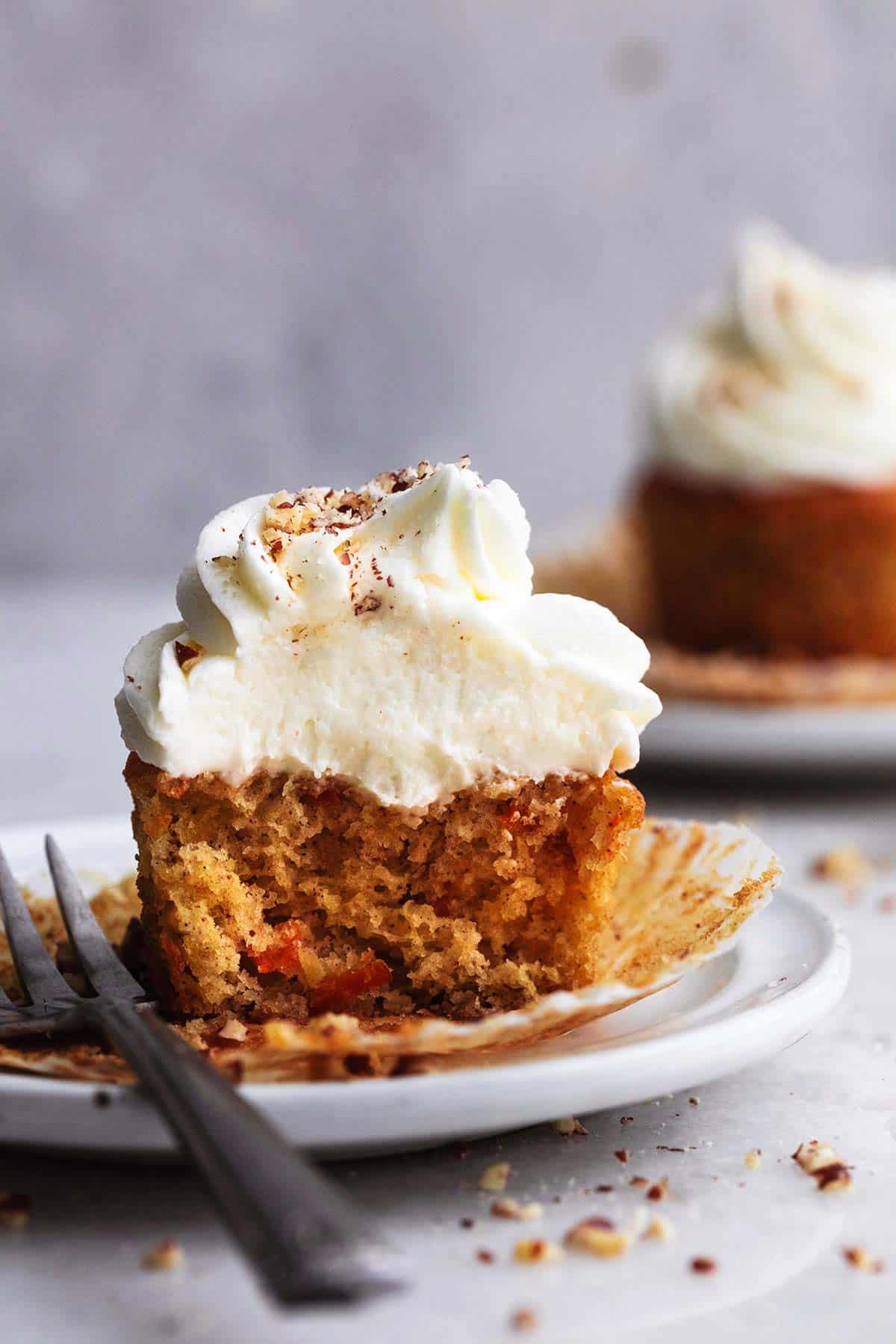 half of a carrot cake cupcake with cream cheese frosting with the liner open with a fork on a plate.