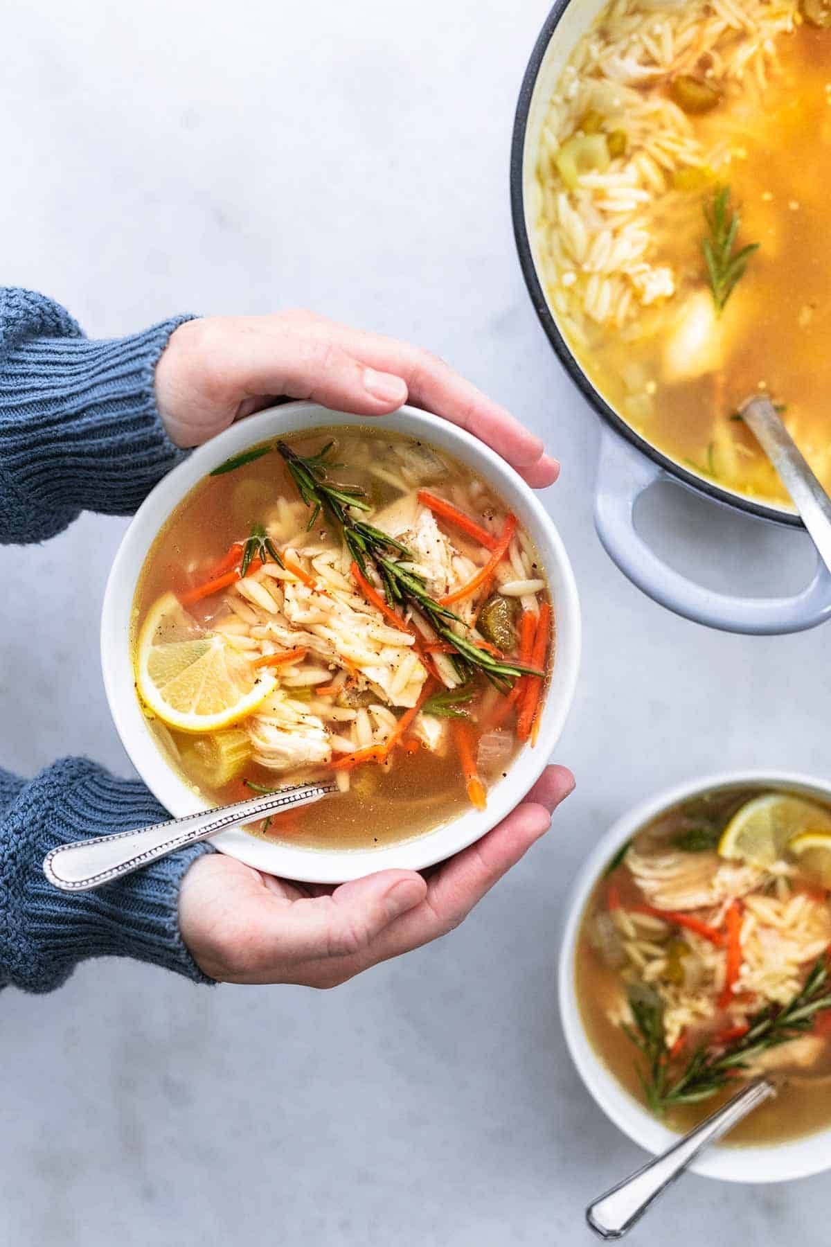 top view of hands holding a bowl of lemon chicken orzo soup with another bowl and pot on the side.