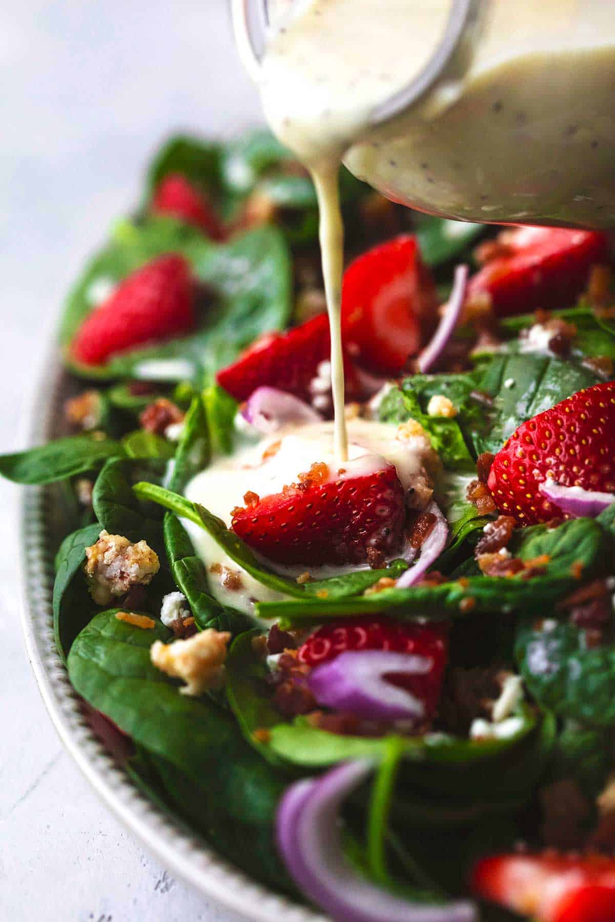 up close view of creamy white salad dressing being poured onto strawberries in spinach salad with red onions and bacon crumbles