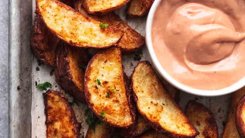 browned crispy potato wedges on baking sheet with pink-brown dipping sauce in white bowl