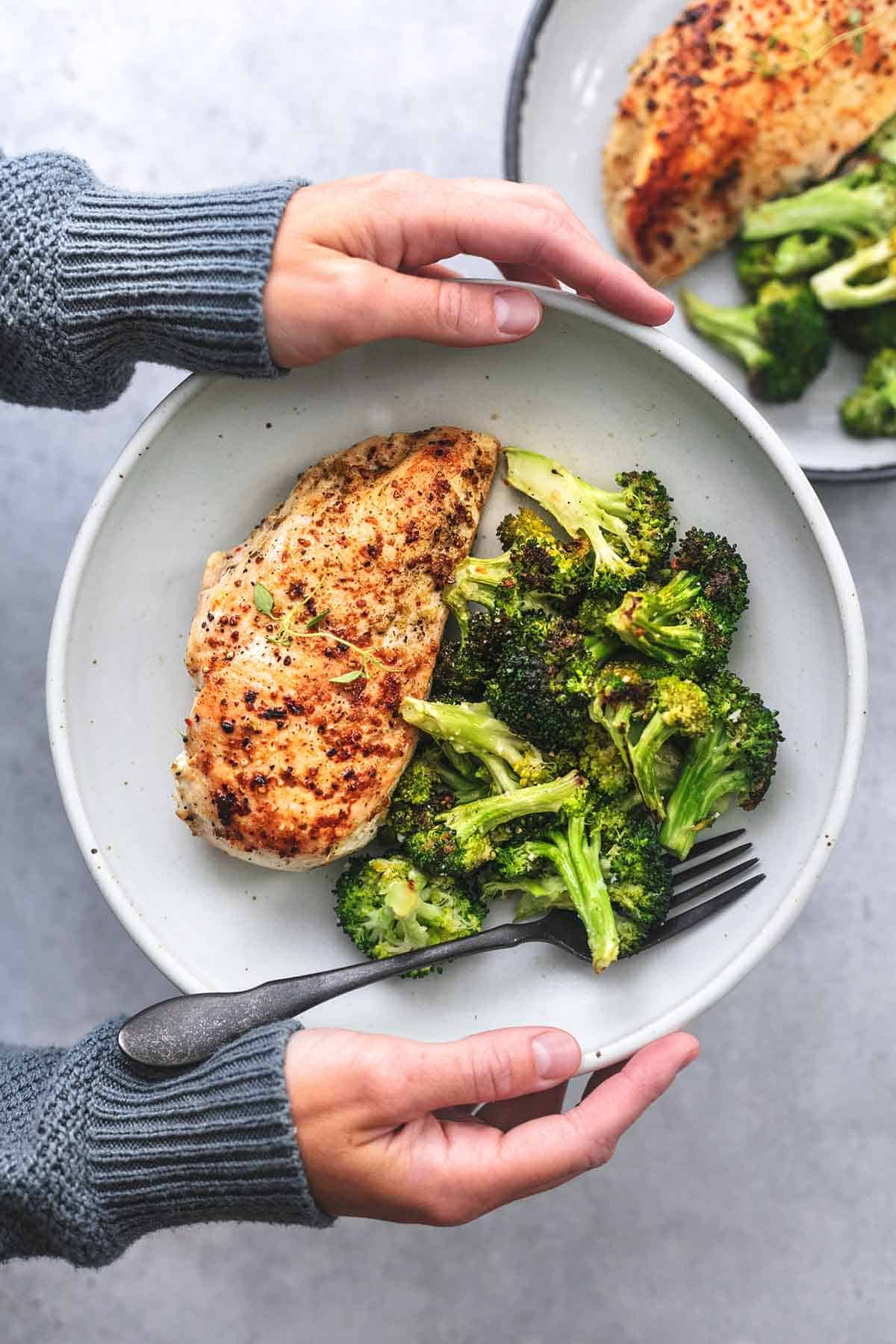 overhead view of hands holding plate of cooked chicken and broccoli above second plate of food