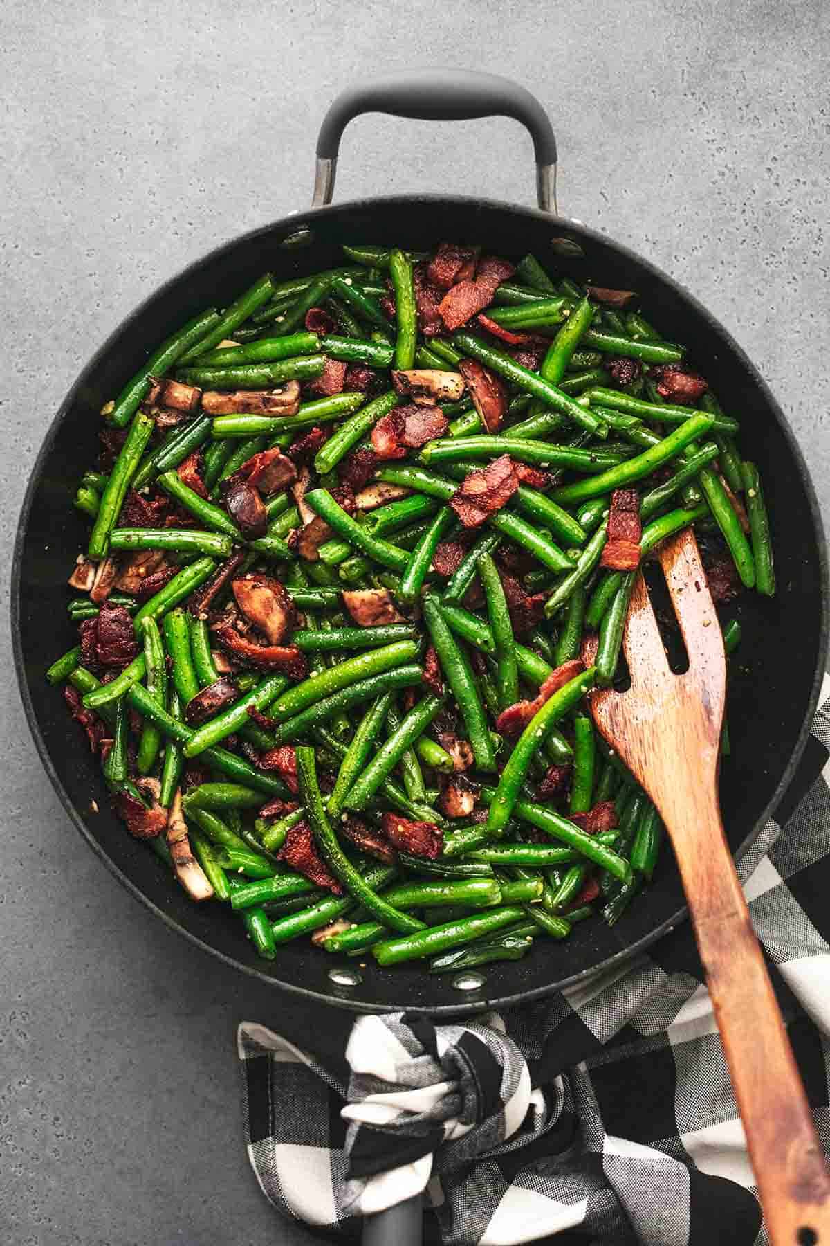 top view of green beans with mushrooms and bacon with a wooden spatula in a skillet.