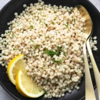 overhead view of cooked pearl couscous with lemon wedges and fresh herbs on black platter