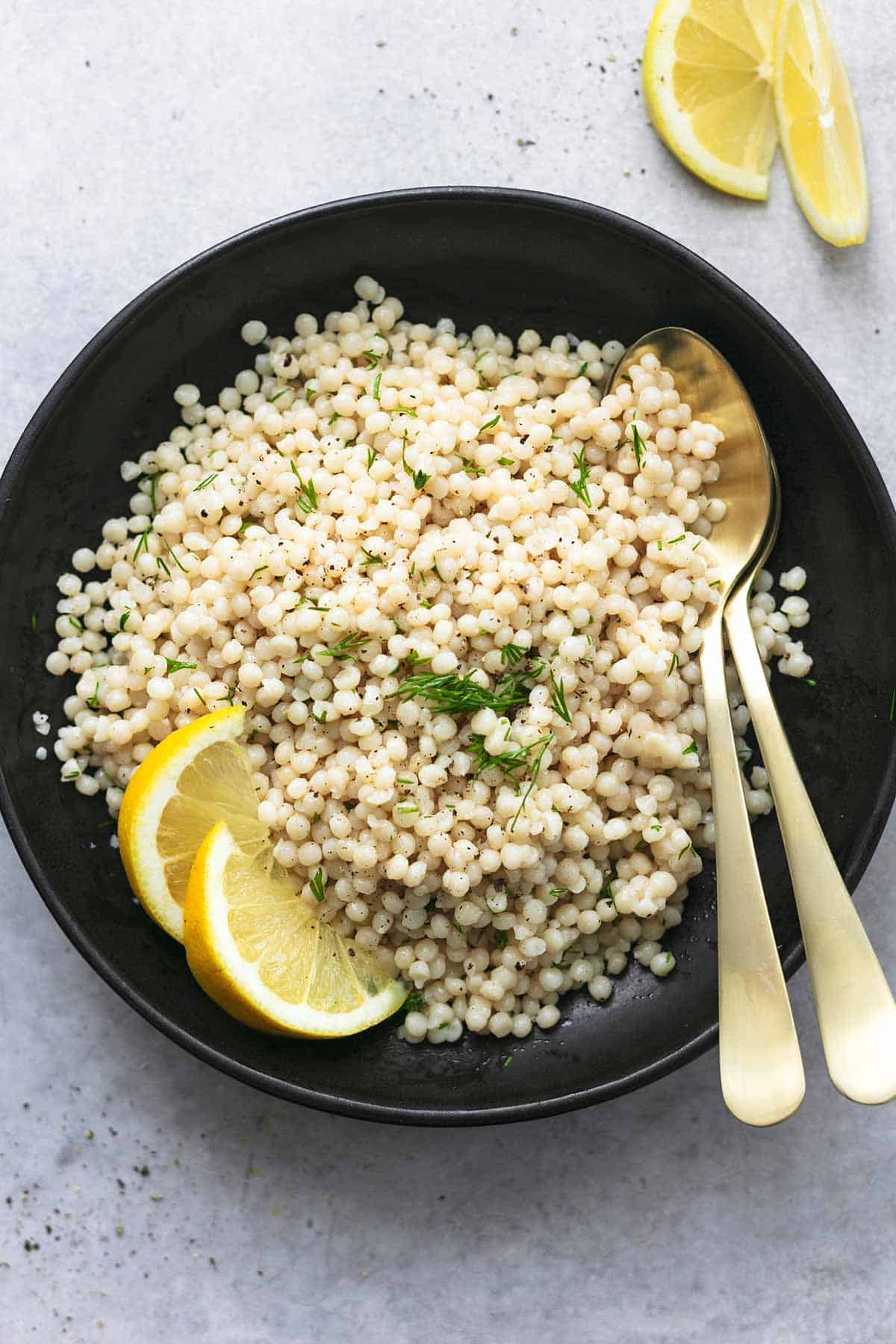 overhead view of cooked pearl couscous with lemon wedges and fresh herbs on black platter