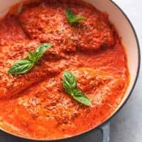 three chicken breasts cooked in red sauce with fresh basil leaves on top in skillet