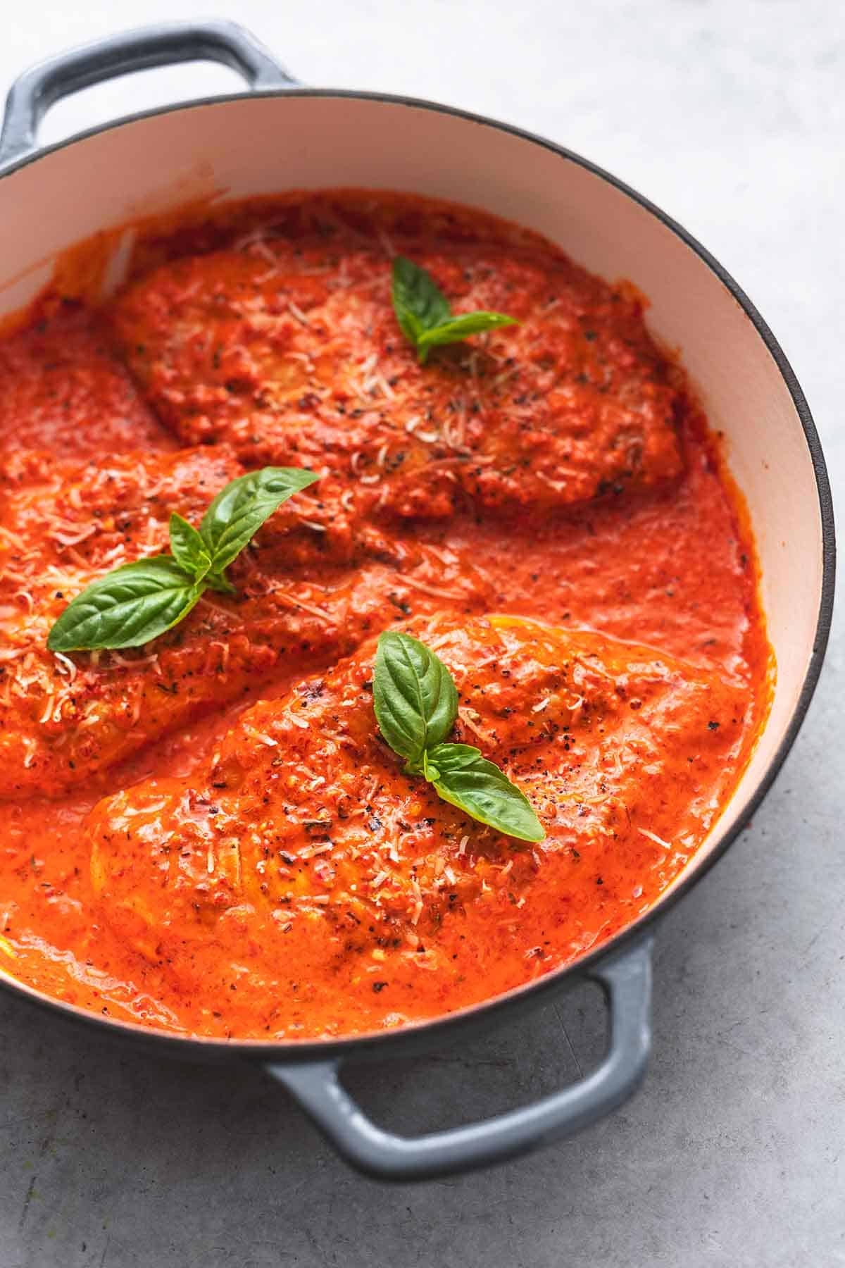 three chicken breasts cooked in red sauce with fresh basil leaves on top in skillet