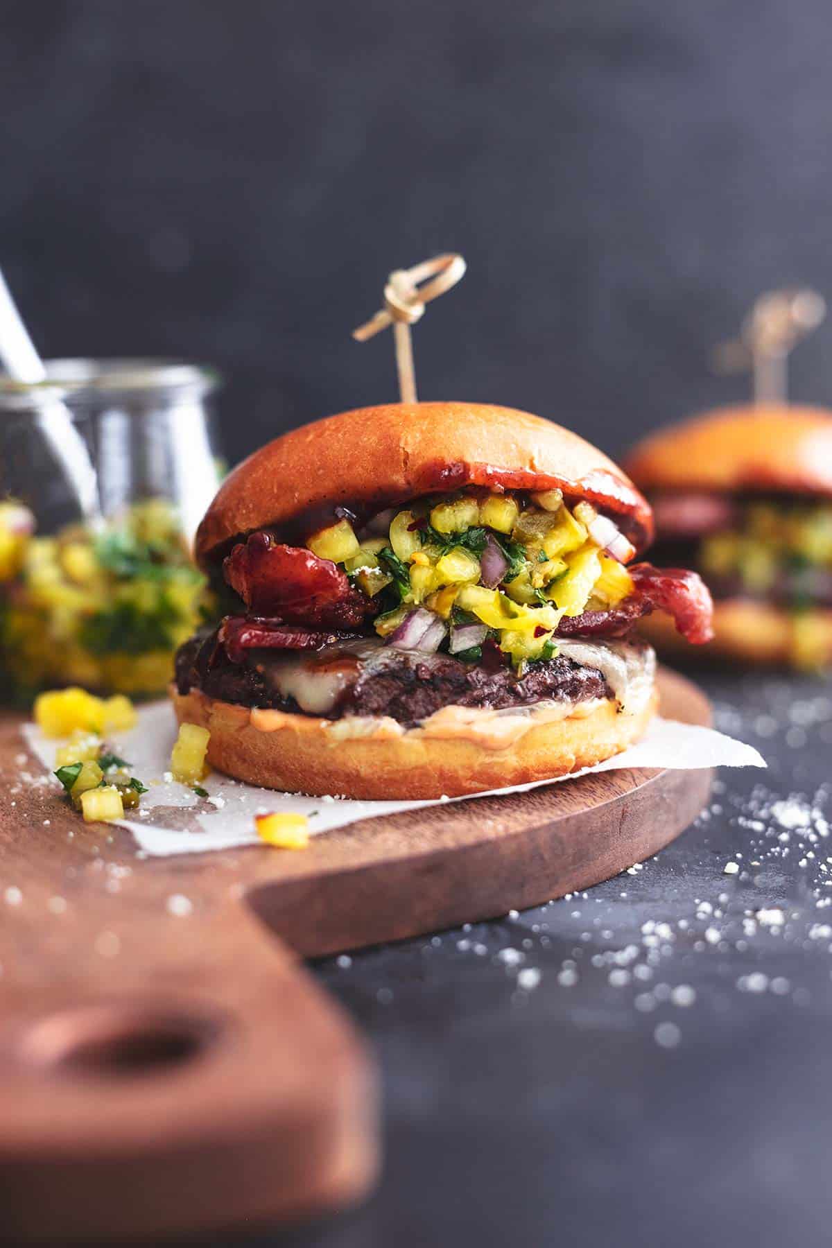 hamburger on a wood cutting board with jar of salsa and second burger behind