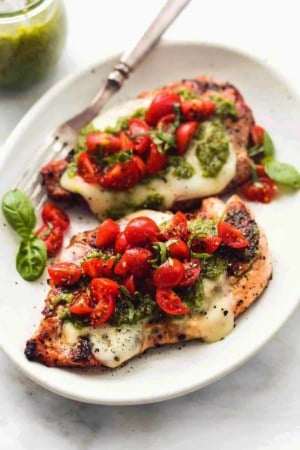 two cooked chicken breasts on a plate, topped with melted cheese, pesto sauce, and cherry tomatoes