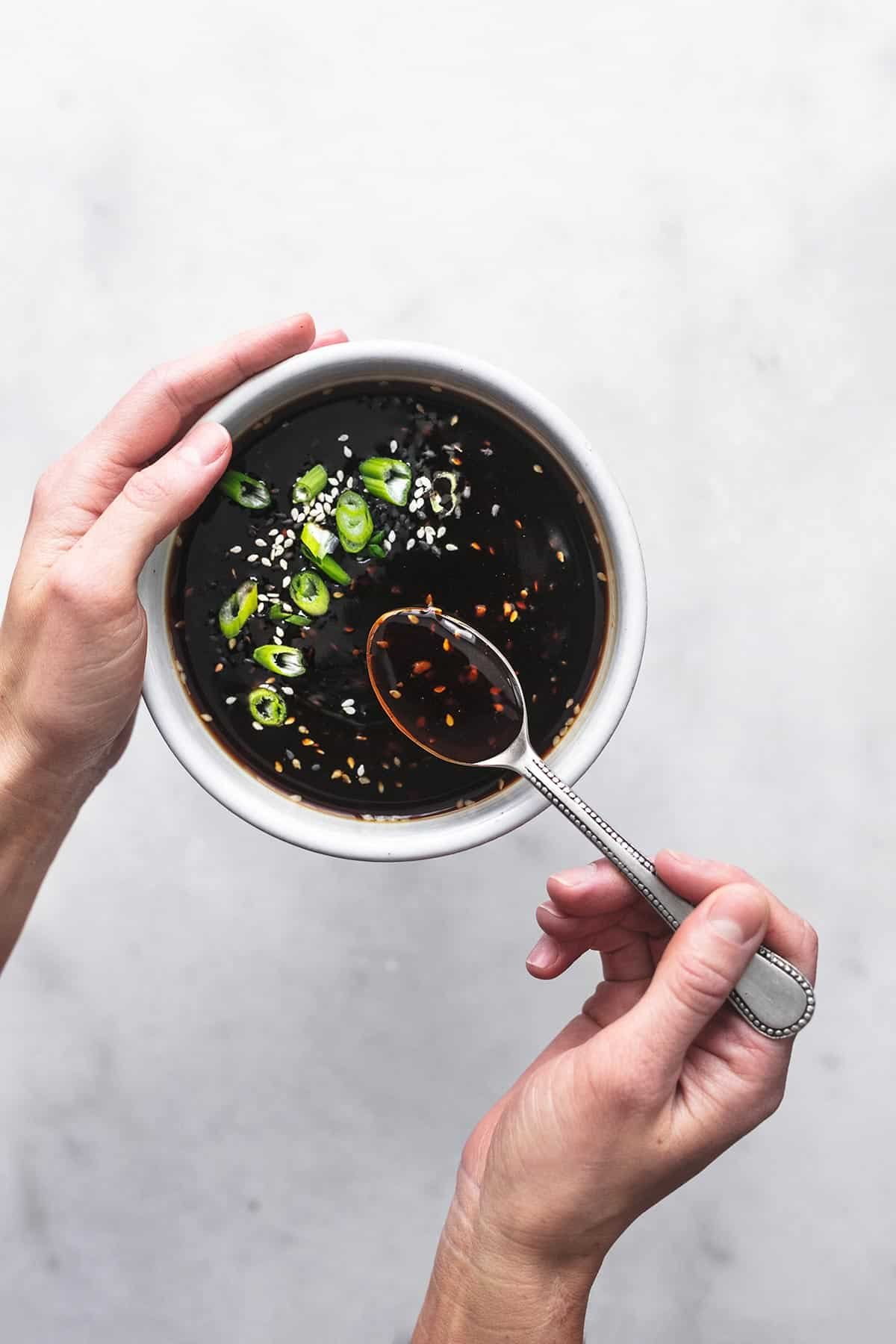 hands holding spoon and bowl of dark sauce with green onions and sesame seeds on top