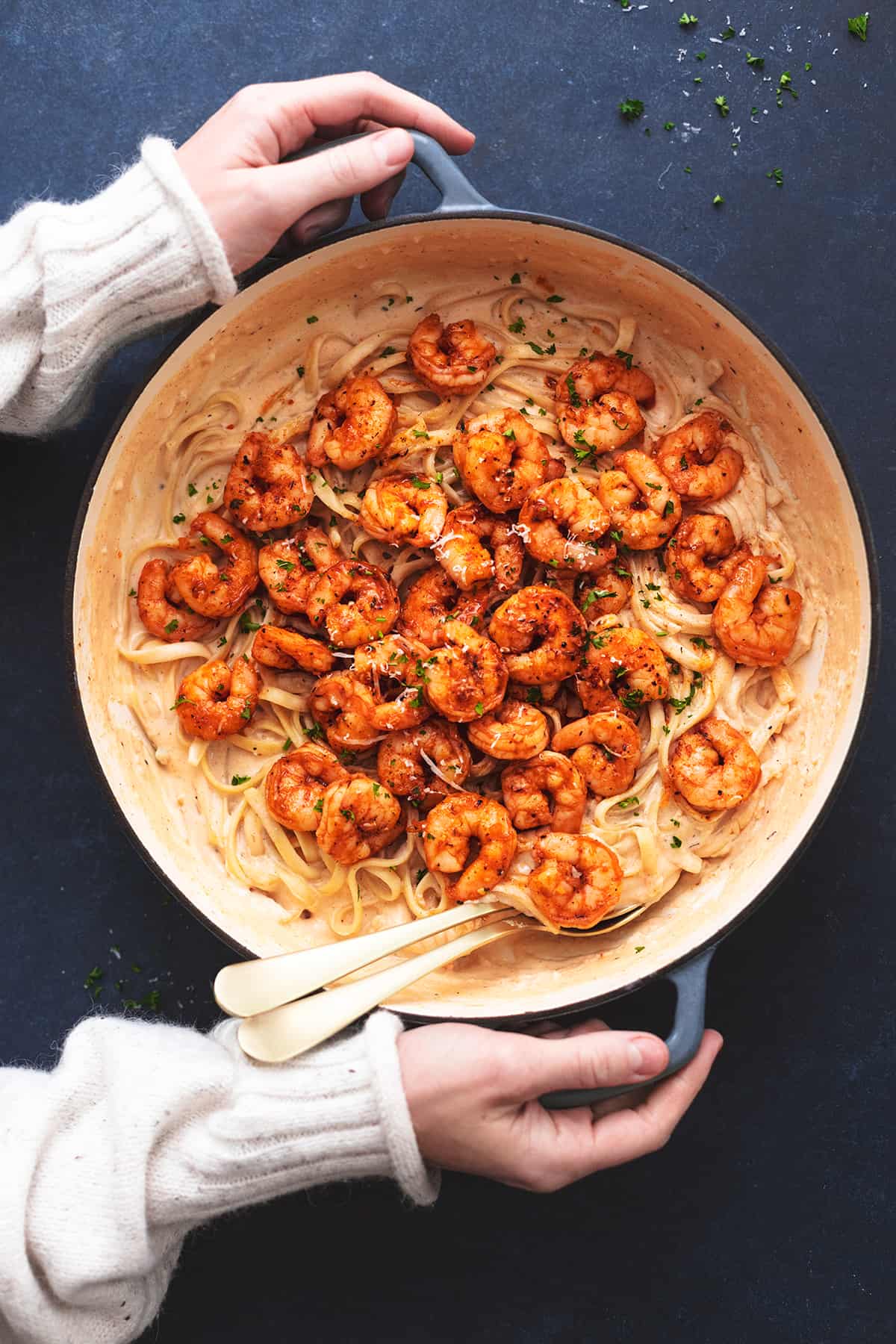 hands holding handles of a skillet filled with pasta noodles and cooked shrimp with two serving spoons