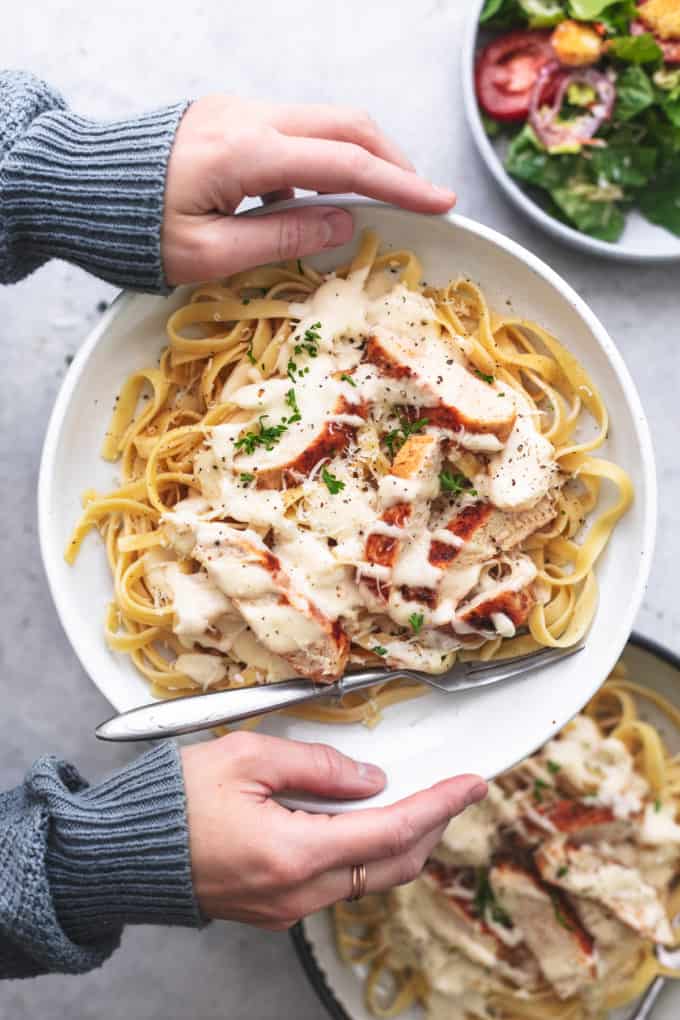 hands holding plate of chicken alfredo pasta above second plate of pasta