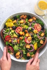 hands tossing salad with wooden spoons and jar of dressing