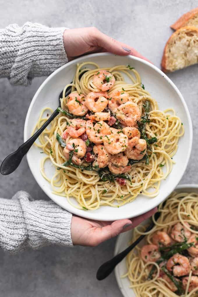 hands holding plate of shrimp and noodles above second plate and piece of bread