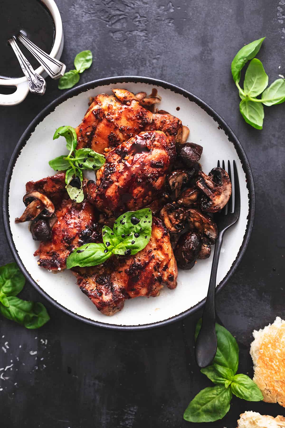 plate of baked chicken thighs with sauce in bowl and basil and bread on table