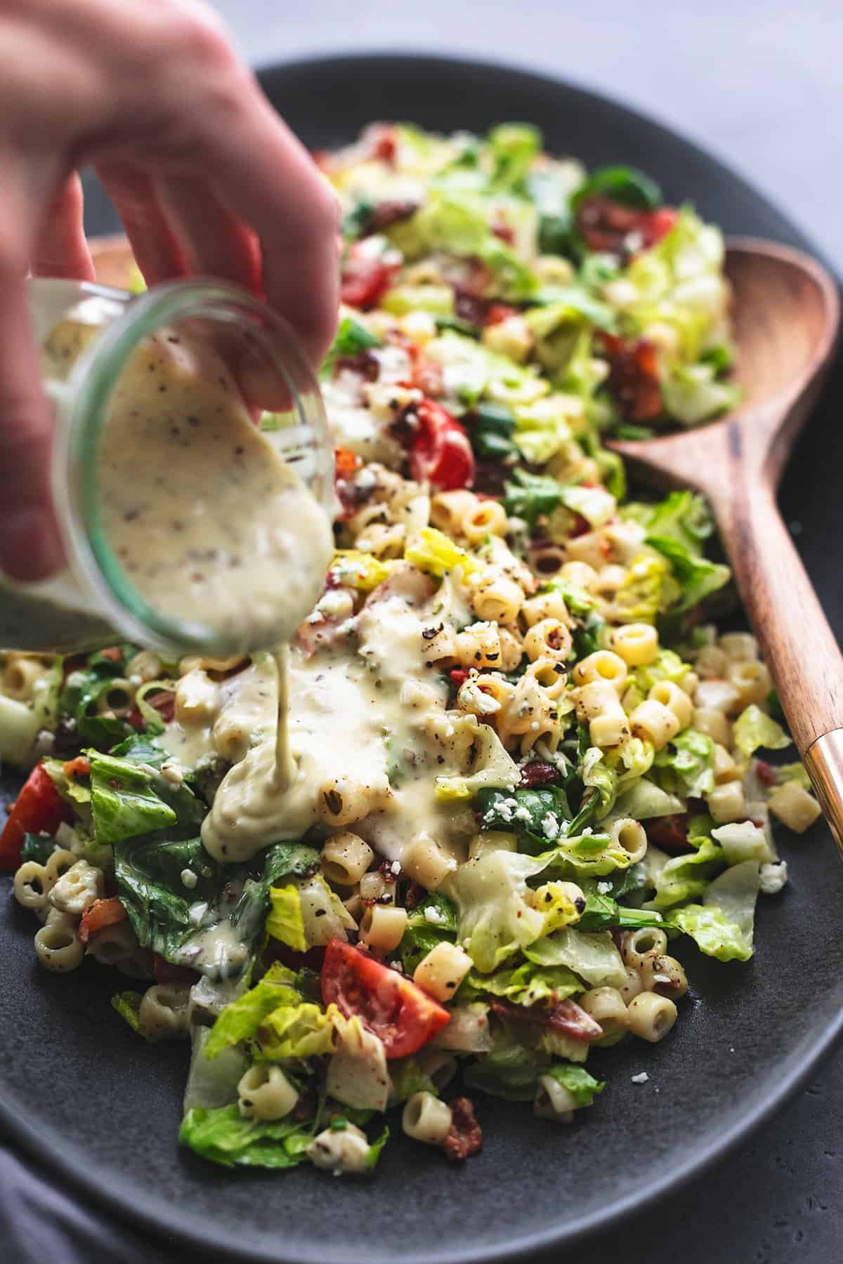 hand pouring dressing over pasta and green salad with tomatoes and bacon