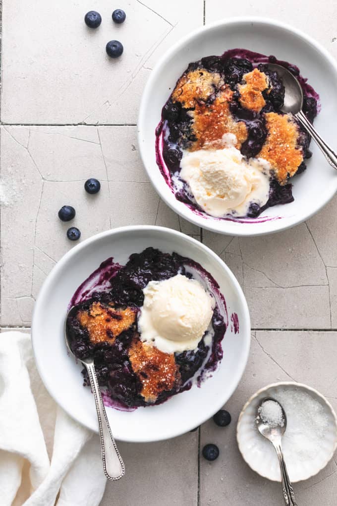 two bowls of blueberry cobbler on a table with napkin and fresh blueberries