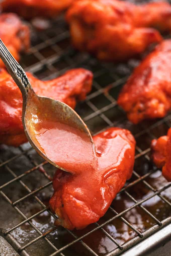 spoon pouring buffalo sauce onto chicken wings on a baking rack