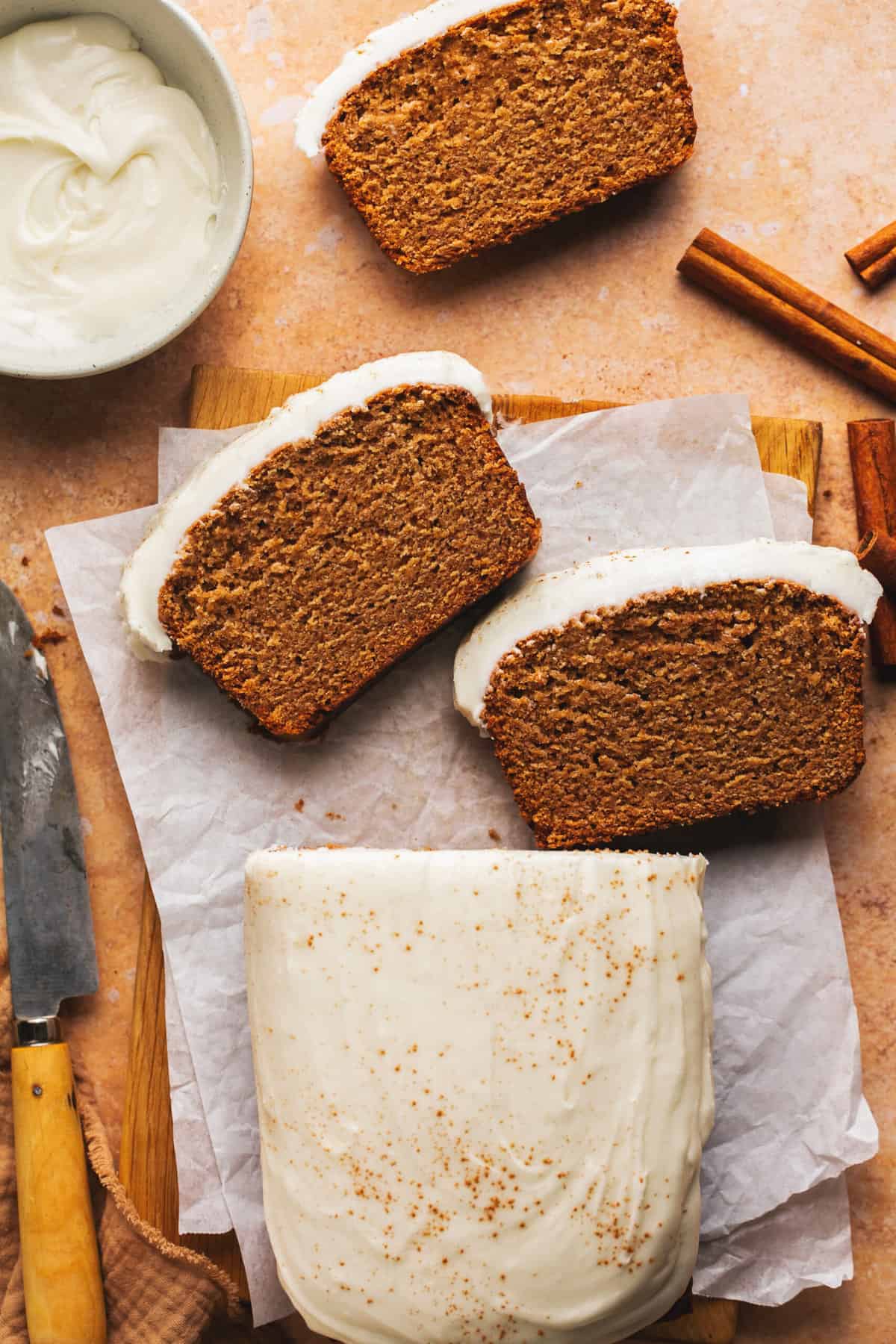sliced loaf of gingerbread with frosting on tabletop with cinnamon sticks and knife