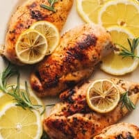 four baked chicken breasts on platter topped with lemon slices and rosemary