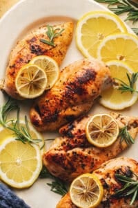 four baked chicken breasts on platter topped with lemon slices and rosemary