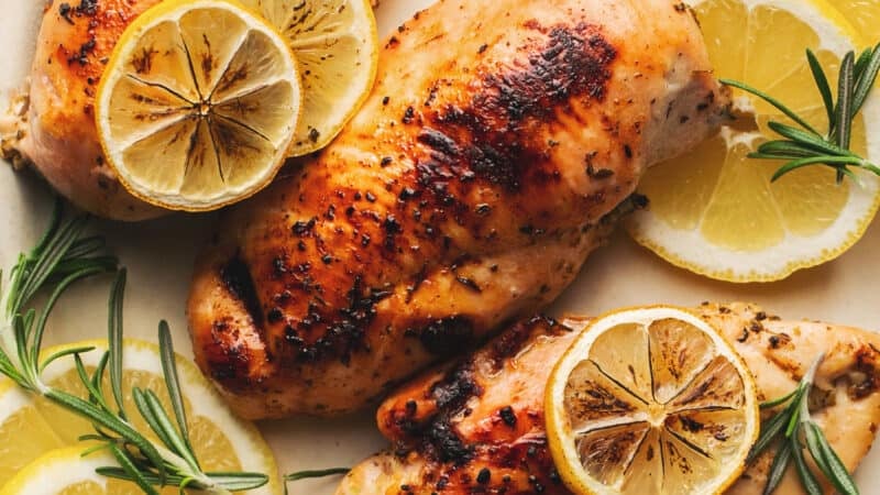 four baked chicken breasts on platter topped with lemon slices and rosemary