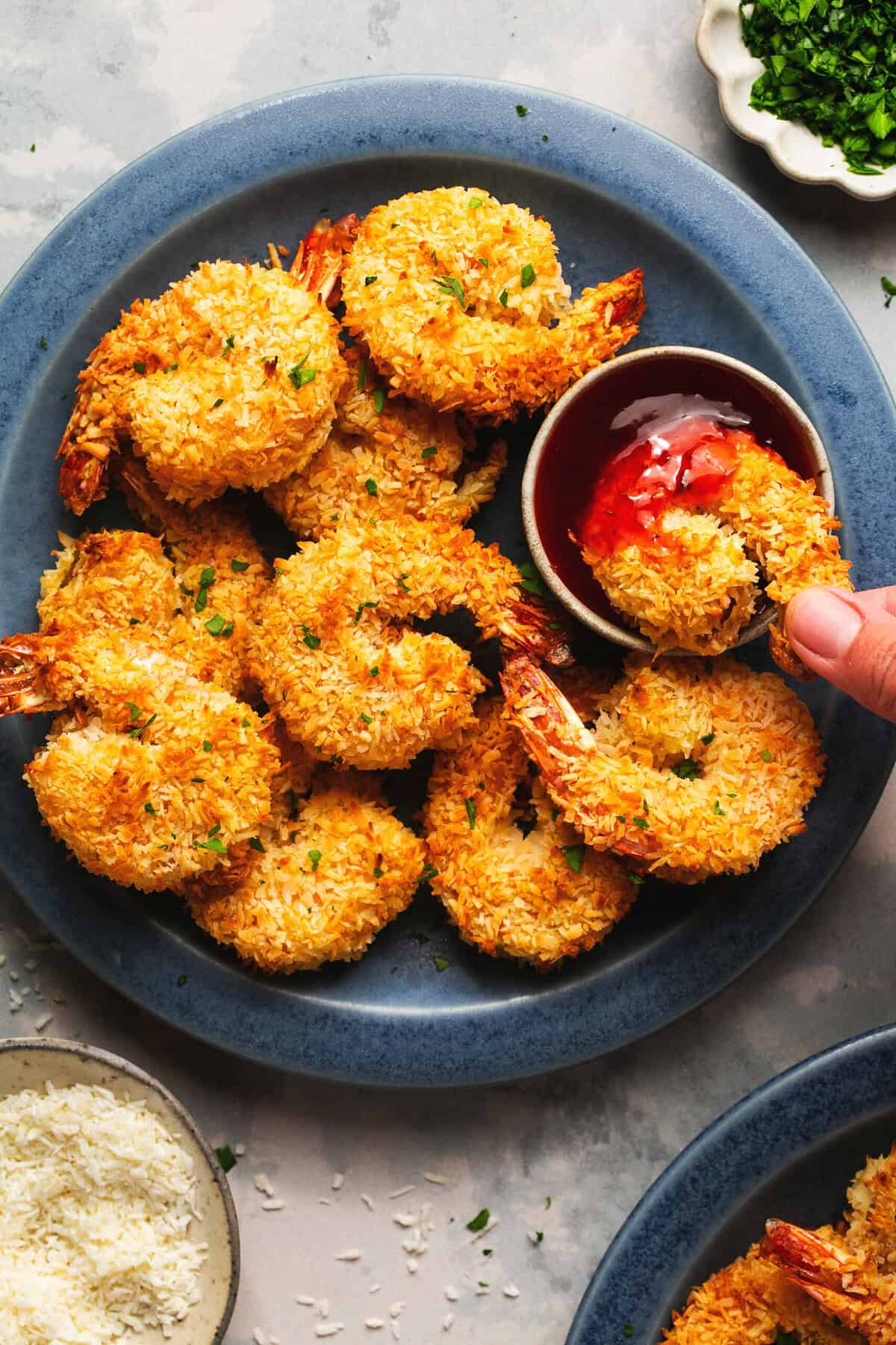 fingers dipping a coconut breaded shrimp into sauce near more shrimp.
