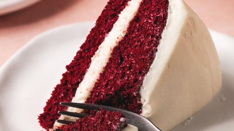 fork cutting into slice of layer cake with cream cheese frosting on plate