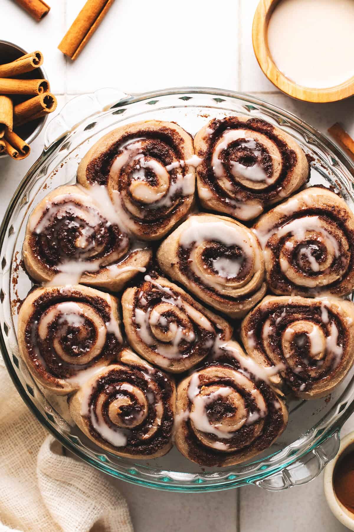 glazed cinnamon rolls on table with napkin, cinnamon sticks, and glaze bowl.