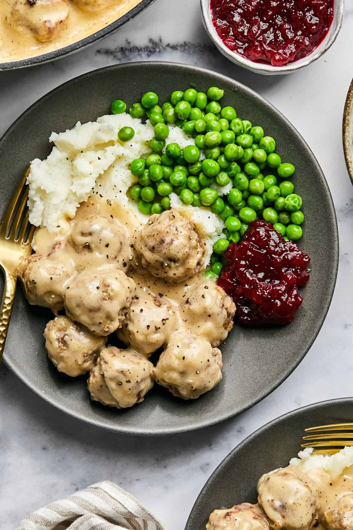 Close up of Swedish meatballs, mashed potatoes, green peas, and lingonberry jam on a grey plate with a fork.