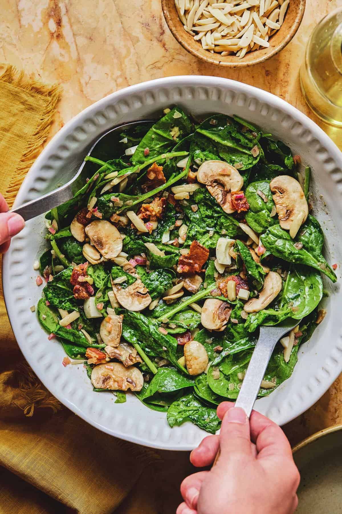 Overhead of two hands holding serving utensils in a dish of spinach salad with hot bacon dressing.